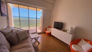 A living room with a sofa, a TV on a white cabinet, and a balcony with a view of the sea.