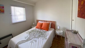 A bedroom with a bed, a nightstand, a desk, a window with blinds, and a door.