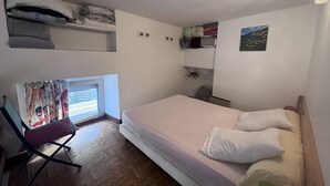 A small, compact room with a single bed, a chair, a window with a view, and a shelf with personal items.