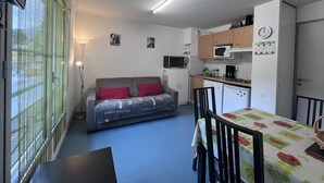 A compact living space with a kitchenette, a sofa with red cushions, a dining table with chairs, and a view through a window with a green curtain.