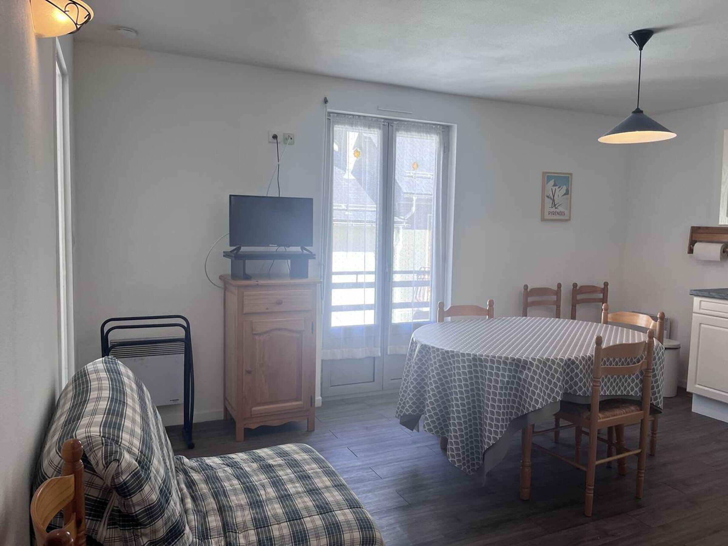 A room with a bed, a TV on a wooden cabinet, a dining table with chairs, and a window with a view of the outside.