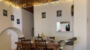 A dining area with a wooden table and chairs, string lights, framed pictures on the wall, and a window with a view.