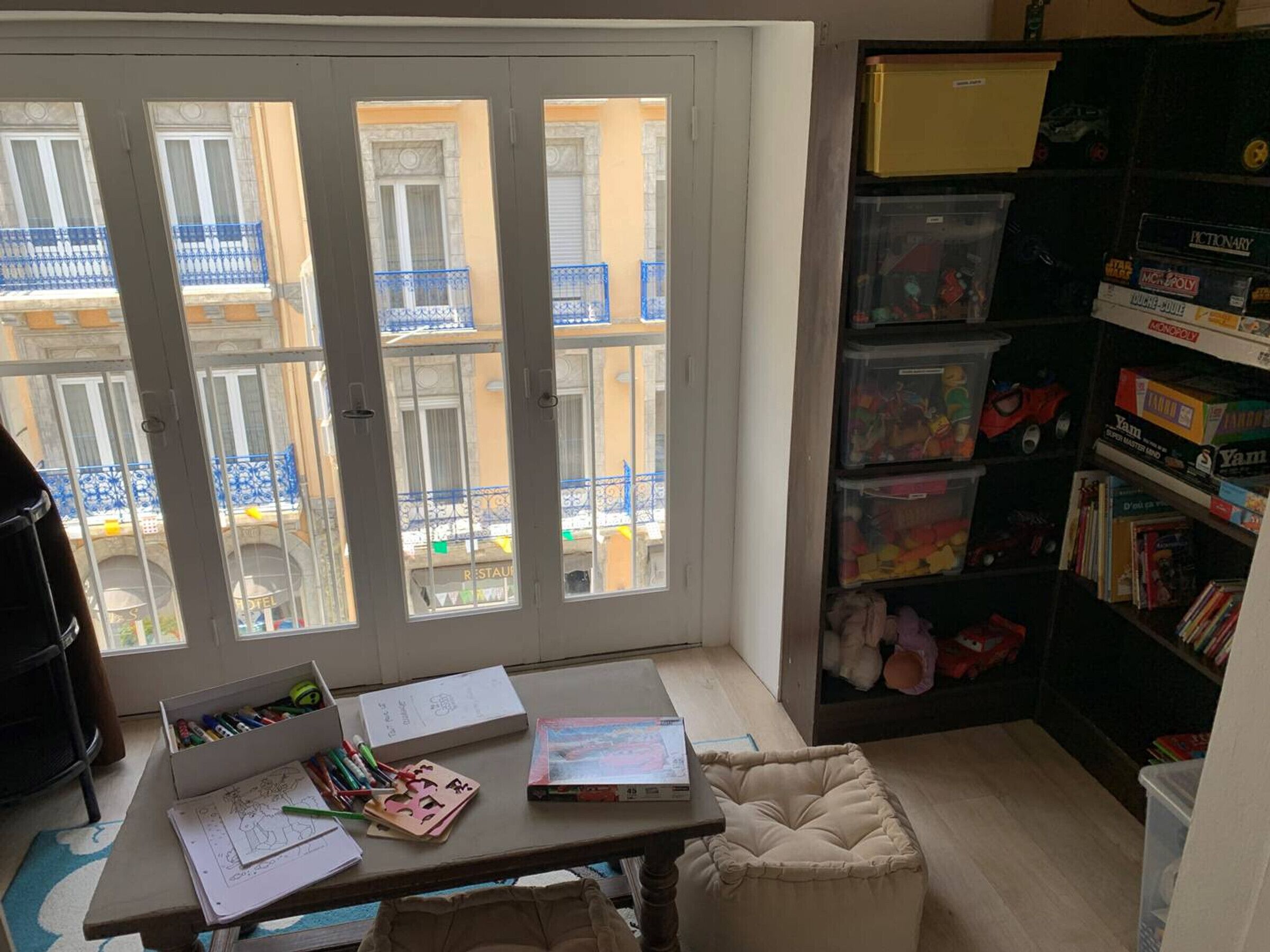 Una habitación con un gran ventanal, un escritorio con material de papelería, una estantería con juegos de mesa y un banco acolchado.