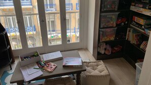 A room with a large window, a desk with stationery, a bookshelf with board games, and a cushioned bench.