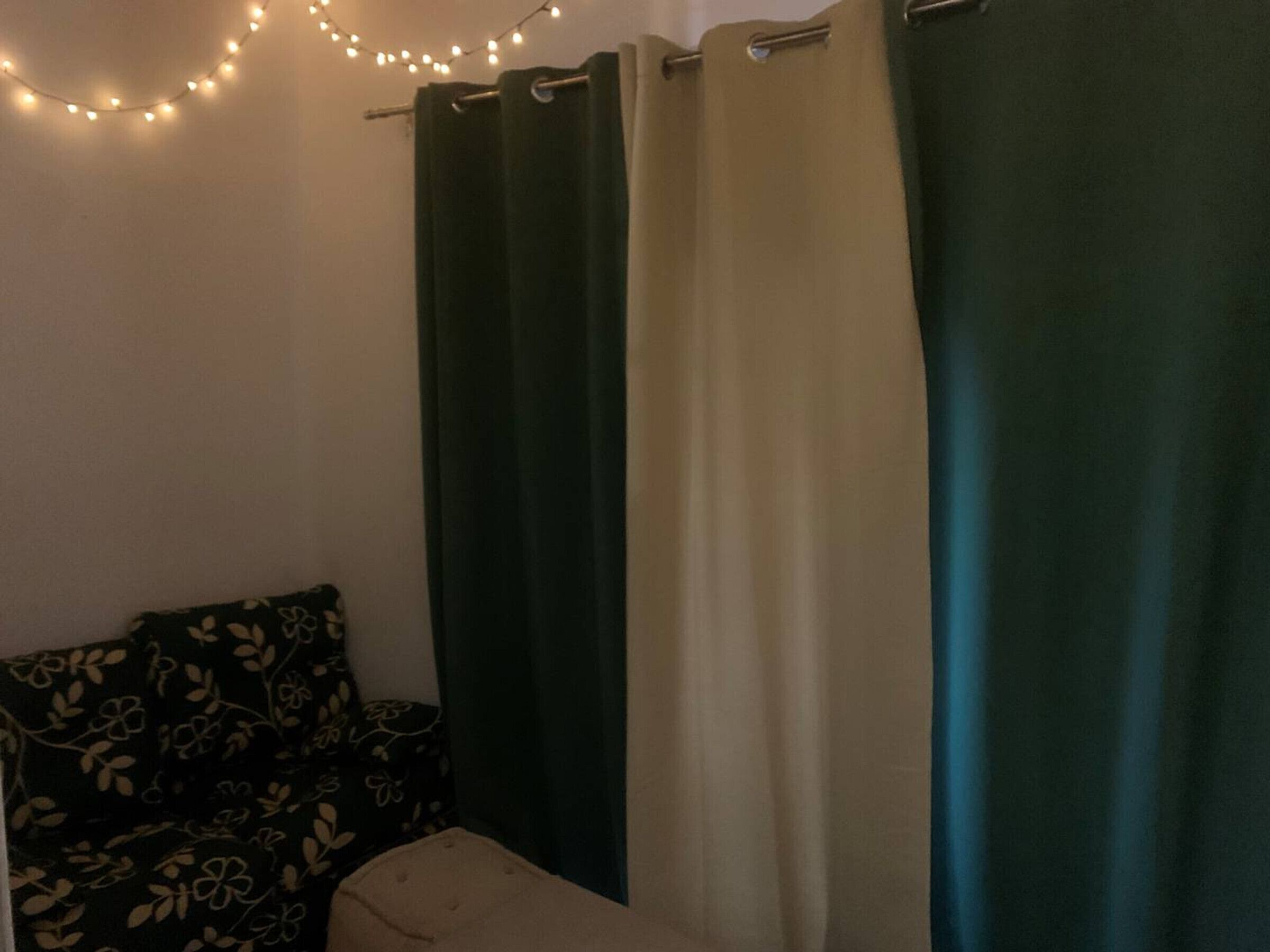 Um quarto com sofá e duas cortinas, uma escura e outra clara, com luzes de cordão acima.
