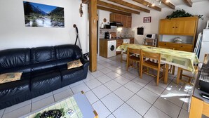 A cozy living room with a black leather sofa, a dining table with chairs, a kitchen area with wooden cabinets, and a large window with a mountain view.