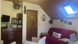 A cozy living room with a wooden ceiling, a red sofa, white chairs, a TV, and a bookshelf.