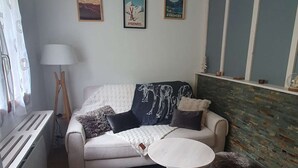 A living room with a sofa, a round table, and two ottomans. There are framed pictures on the wall, a lamp, and a radiator. - Charming chalet for 4 in Cauterets with parking (Cauterets)