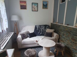 Uma sala de estar com sofá, mesa redonda e dois banquinhos. Há quadros nas paredes, um abajur e um radiador.