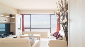 A modern living room with a large flat-screen TV, a sofa, a coffee table, and a view of the sea through large windows.