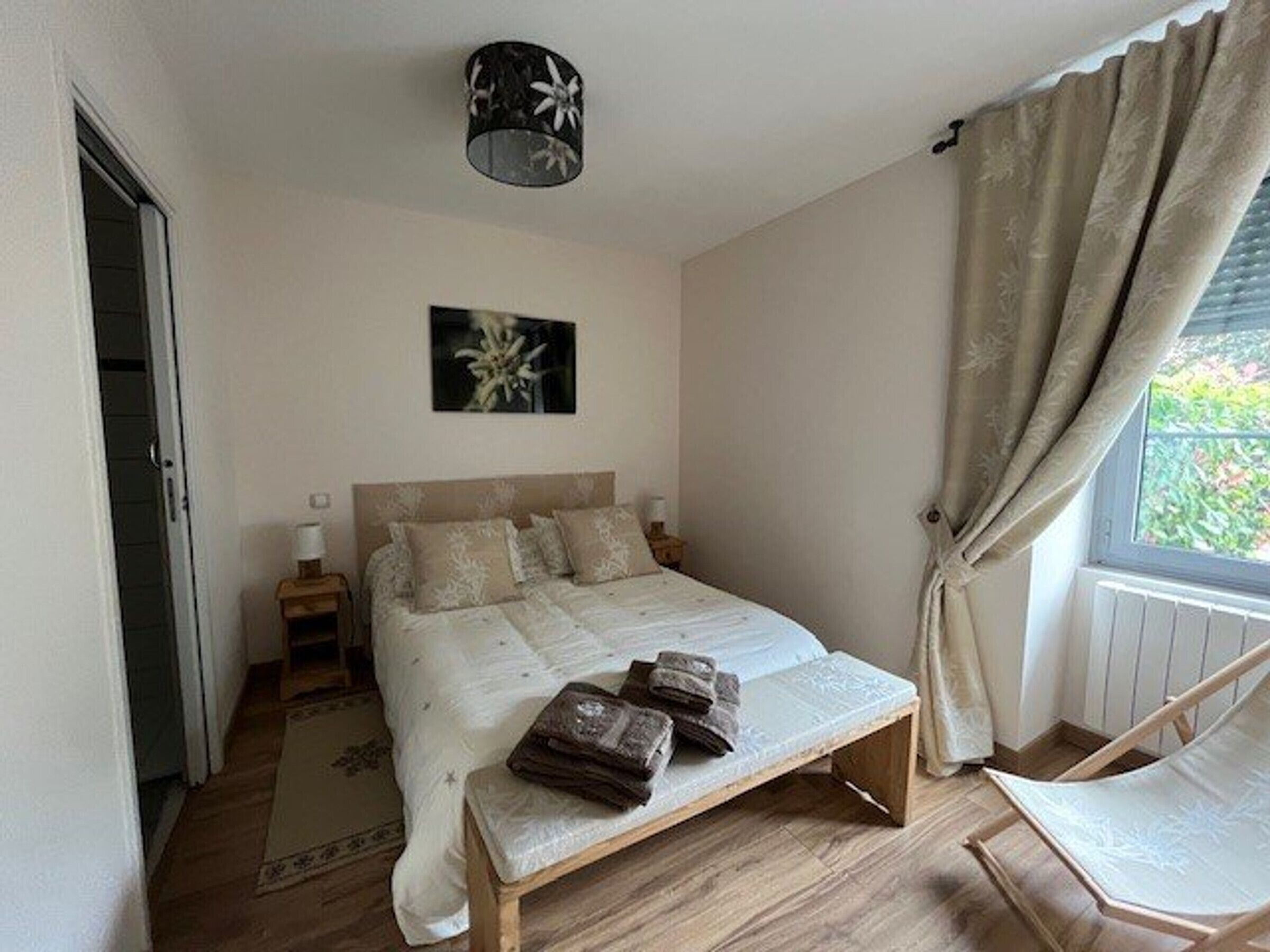 Ein Schlafzimmer mit Bett, Nachttischen, einer Bank, einem Sessel, einem Fenster mit Vorhängen und einer Deckenleuchte.