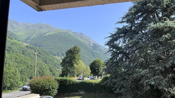 A balcony with a view of a mountain range, a parking area with cars, and a large evergreen tree.