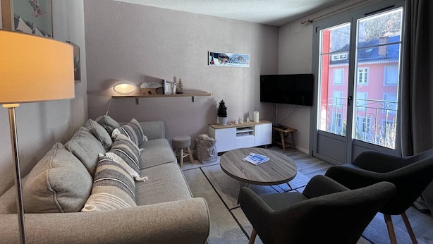 Ett modernt vardagsrum med en soffa, fåtöljer, ett soffbord, en tv och ett fönster med utsikt över byggnader.