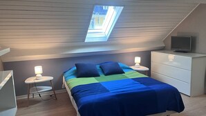 Ett sovrum med ett sluttande tak, ett takfönster, en säng med ett blått och grönt sängöverkast, två sänglampor, en tv på en byrå och ett litet vitt nattduksbord.