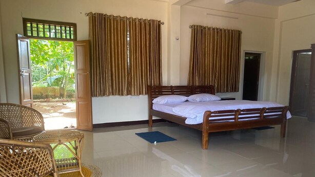 A bedroom with a wooden bed, two wicker chairs, a small rug, and a view of greenery outside. - Kappils INN HOMESTAY (Changanassery)