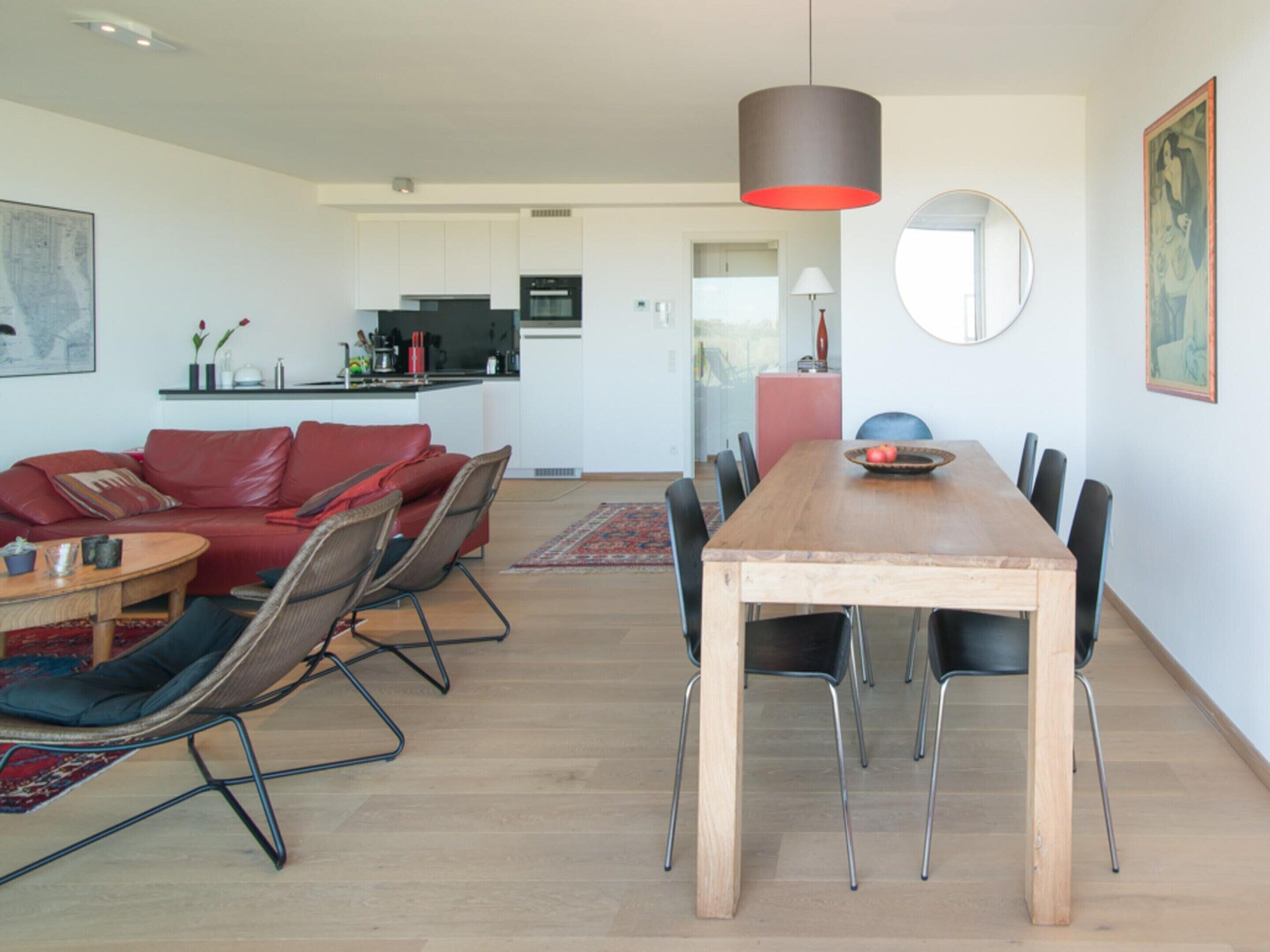 A modern living room with a wooden dining table, red leather sofa, and a kitchen in the background.