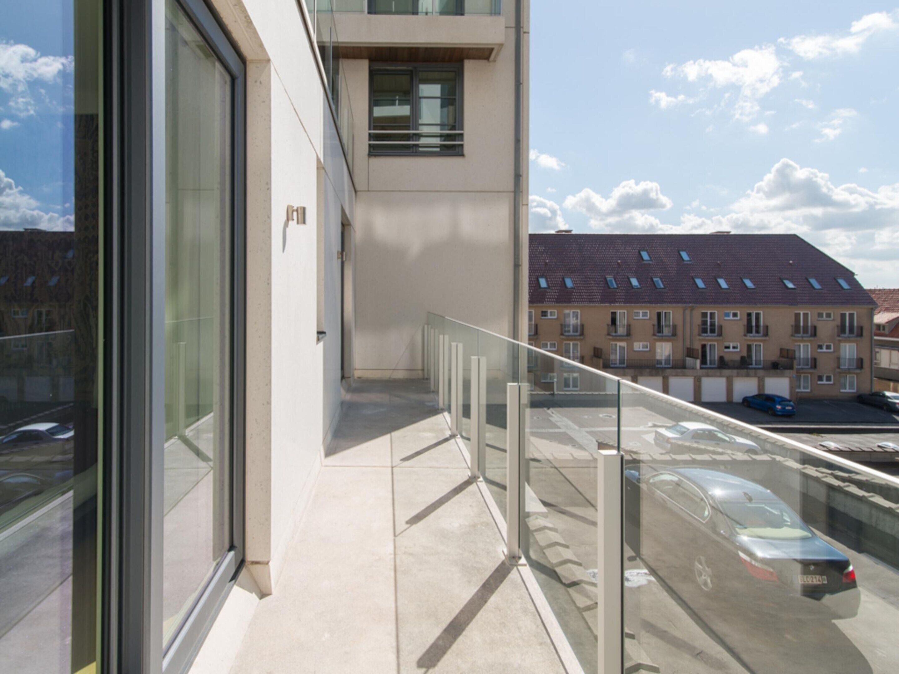 A modern building with a glass balcony, a clear sky, and a residential area with parked cars.