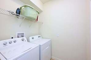 Une buanderie équipée de deux machines à laver et deux sèche-linge superposés, d’une étagère avec un panier, et d’une prise de courant au mur.