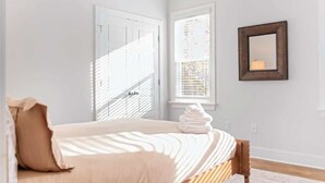 A bedroom with a bed, a window with blinds, a wall-mounted mirror, and a stack of towels. - Family-Friendly 4BR w/ Fenced Yard & Parking (Durham)