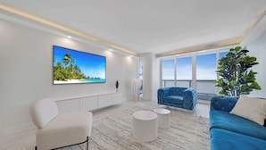 Un salon moderne avec une grande télévision, un canapé bleu, un fauteuil blanc et une grande fenêtre donnant sur la mer.