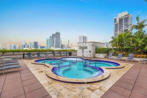 Piscina no terraço com bordas azuis, rodeada por espreguiçadeiras e um horizonte urbano ao fundo.