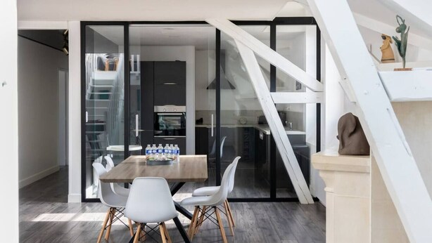 A modern dining area with a wooden table and white chairs, a glass partition, and a staircase. - Artists' Loft (Sainte-Savine)