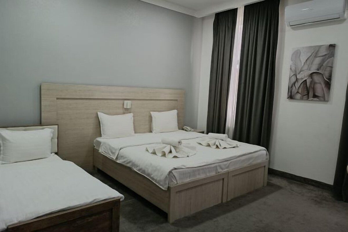 A hotel room with a wooden headboard bed, a smaller bed, a bedside table, a wall-mounted air conditioner, and a painting on the wall.