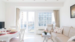 Un salon lumineux avec un canapé blanc, une table à manger dressée avec des assiettes et des verres, et une vue sur le paysage urbain à travers de grandes fenêtres.