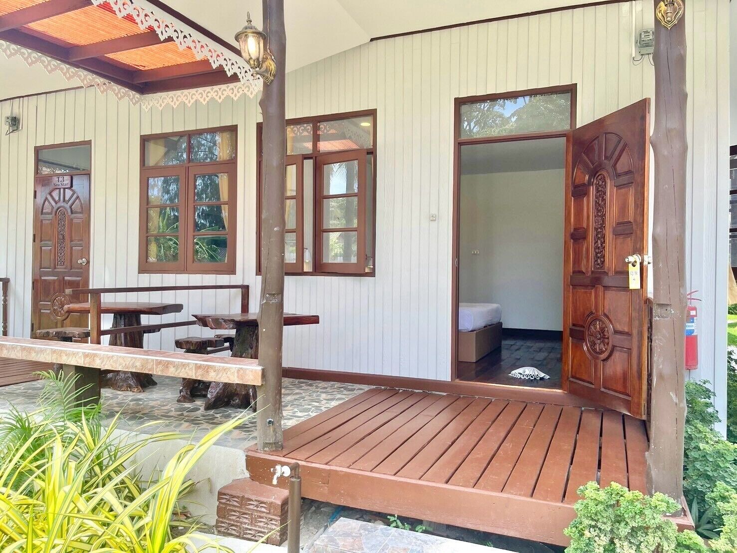 Un porche de madera con un banco y una mesa, una puerta que da a una habitación y una pequeña zona de jardín con plantas.