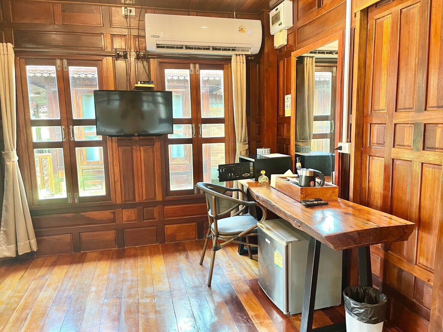 A compact room with a wooden floor, a small desk with a chair, a television, a refrigerator, and a window with curtains.