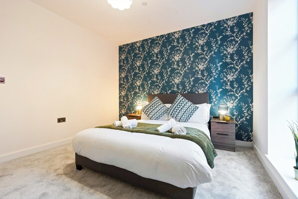 A neatly made bed with white linens, a dark headboard, and a green throw blanket. There is a nightstand with a lamp and a small plant beside the bed. The wall behind the bed has a floral wallpaper pattern. - Camden Drive City Apartment (Birmingham)