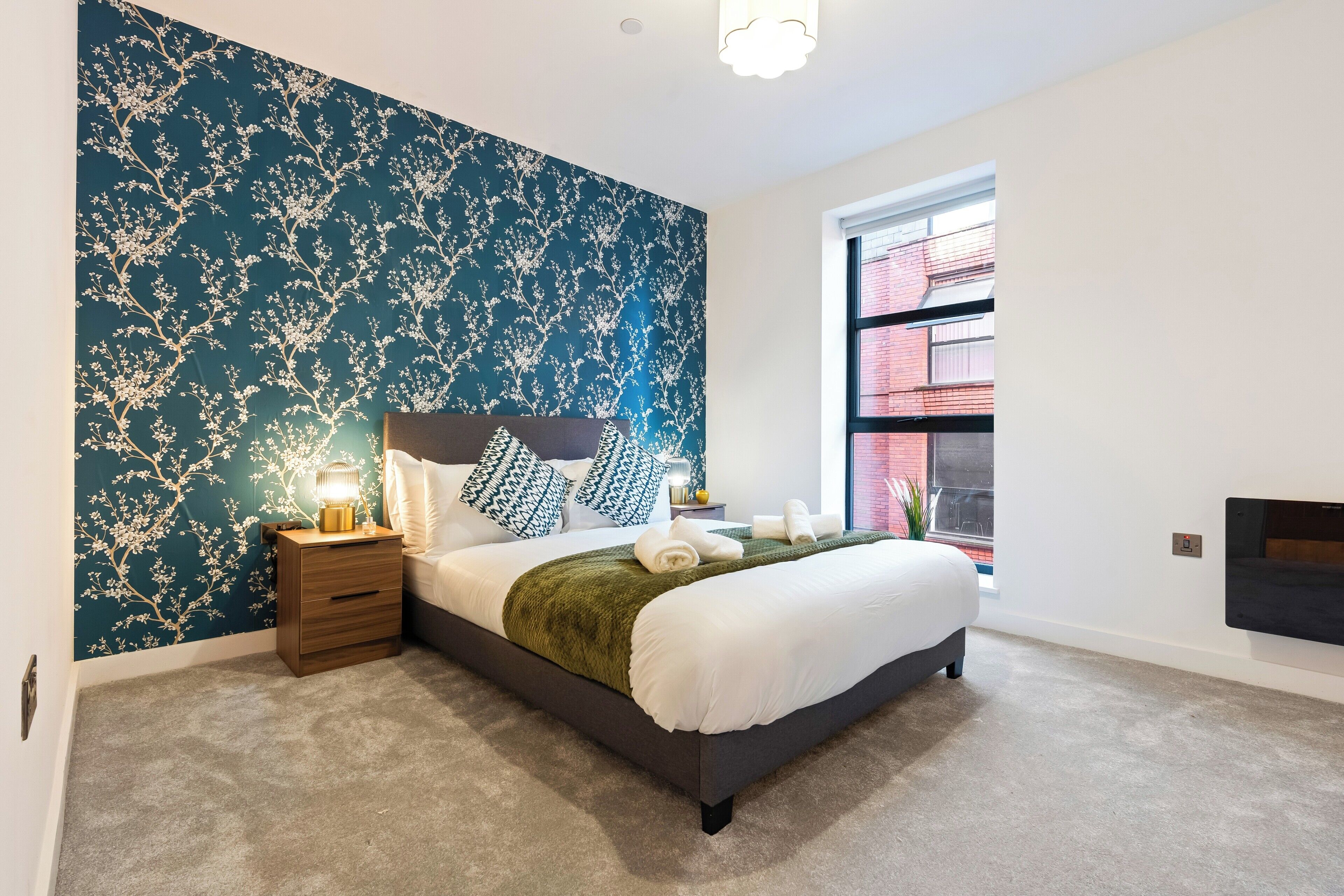 Een slaapkamer met een groot bed, een nachtkastje, een aan de muur bevestigde televisie en een raam met uitzicht op een bakstenen gebouw.