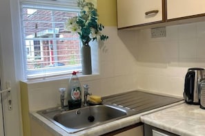 A kitchen with a stainless steel sink, a window with a view of buildings, a vase with flowers, and a kettle on the counter. - Cosy 1 Bedroom Apartment Private Access in Guildford

 (Guildford)