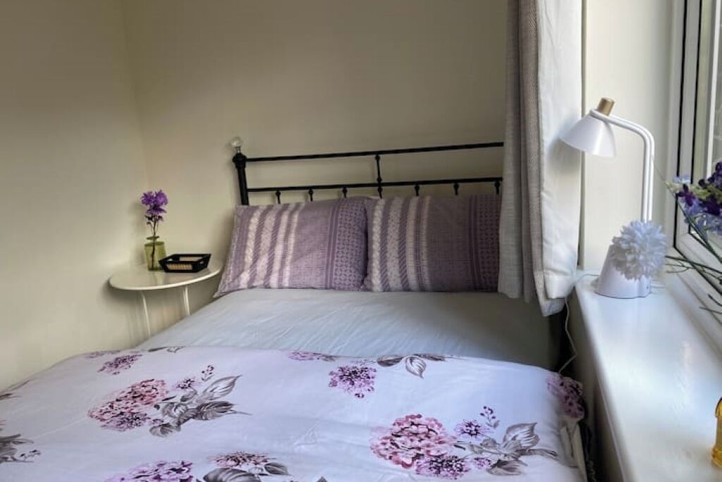 A neatly made bed with floral bedding, a bedside lamp, and a small table with a vase of flowers.
