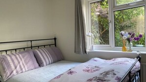 A bed with a floral bedspread, a window with curtains, and a vase with flowers on a windowsill. - Cosy 1 Bedroom Apartment Private Access in Guildford

 (Guildford)