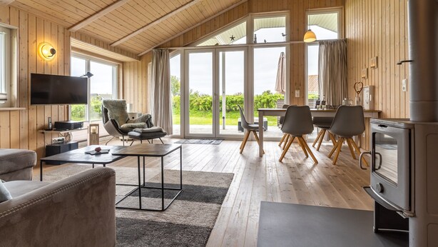A spacious living area with a large TV, comfortable seating, a dining table, and a kitchen with a stove. - Holiday Home by North Sea With Sauna (Wesselburenerkoog)