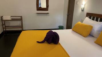 Hotellihuone, jossa on sänky, puinen penkki, peili ja vihreä roskakori.
