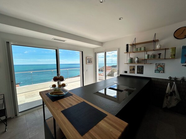 A modern kitchen with a large island, a view of the ocean, and a dining area with a table and chairs. - Dream vacation in a beautiful villa with exceptional sea view (Cerbère)