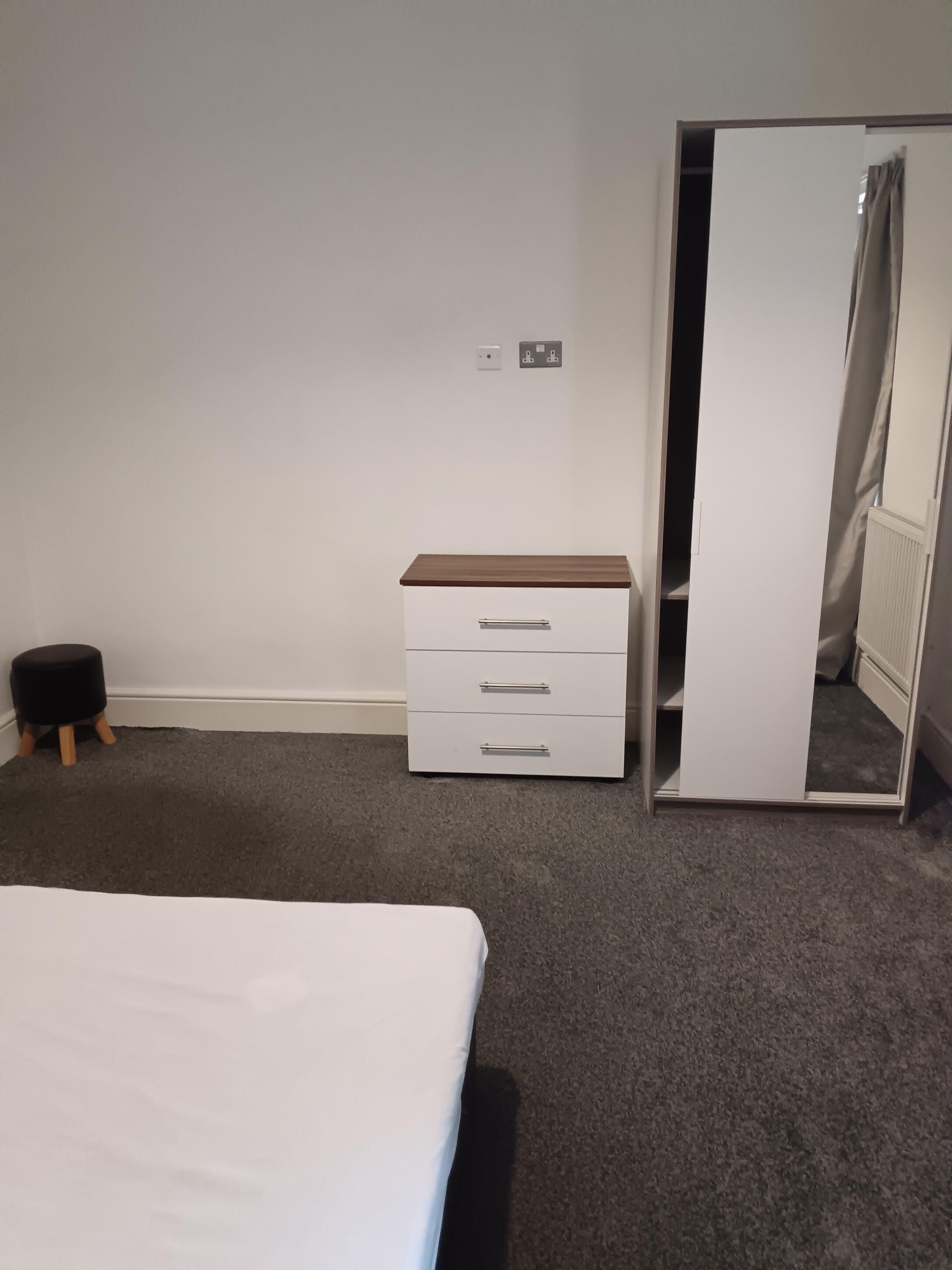 A bedroom with a bed, a white dresser with drawers, a small stool, and a wardrobe with a mirror.
