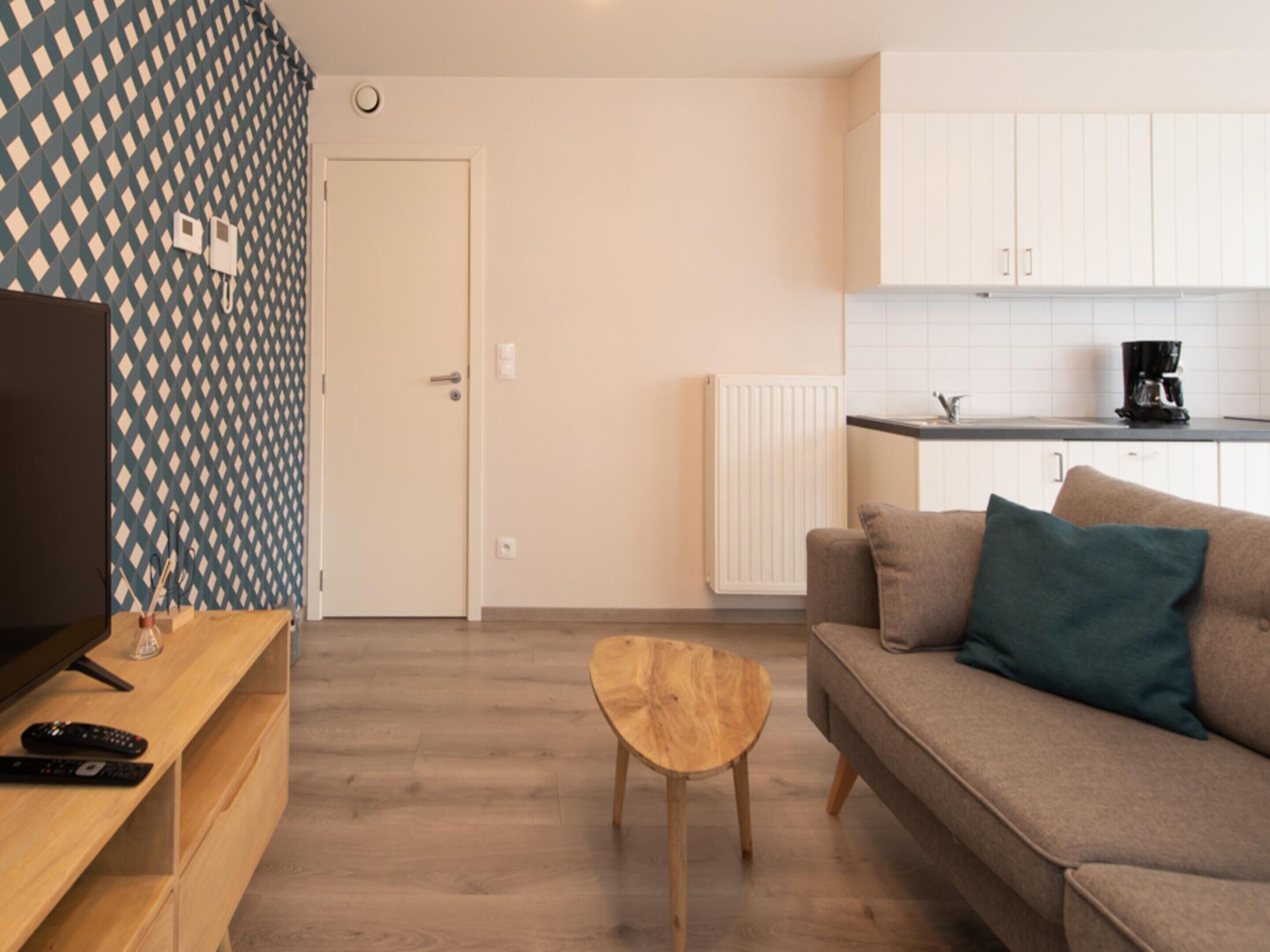 Una sala moderna con un mueble bajo para televisor de madera, una pequeña mesa redonda de madera, un sofá color beige con un cojín azul verdoso, y una zona de cocina con armarios blancos y una cafetera negra.