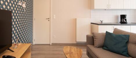 Una sala moderna con un mueble bajo para televisor de madera, una pequeña mesa redonda de madera, un sofá color beige con un cojín azul verdoso, y una zona de cocina con armarios blancos y una cafetera negra.