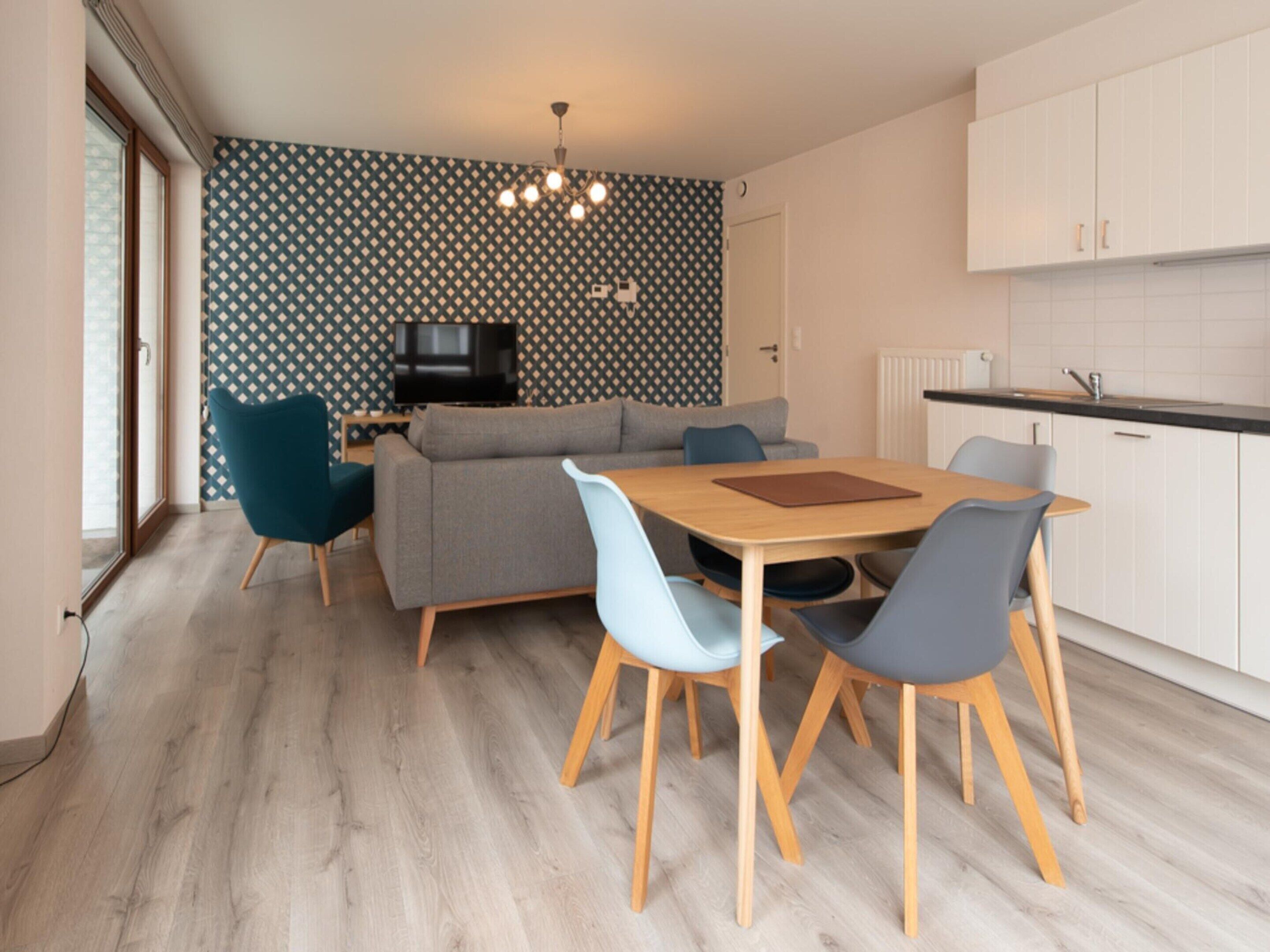 A modern kitchen with white cabinets, a dining table with chairs, a grey sofa, and a TV on the wall.
