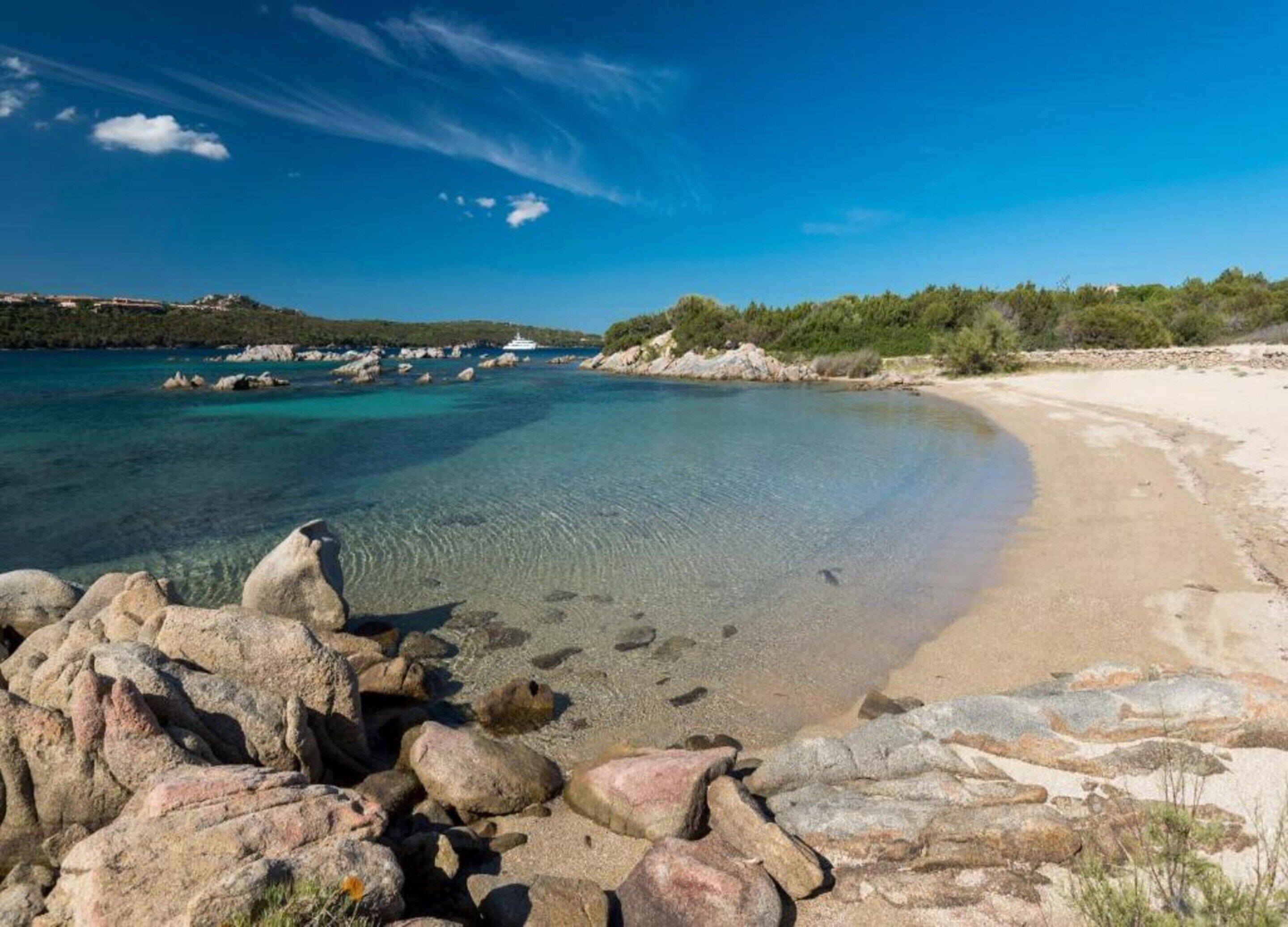 Una spiaggia con acque cristalline turchesi, un litorale roccioso e edifici in lontananza.
