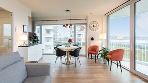 A modern living room with a grey sofa, a dining table with chairs, and a large window offering a view of buildings and a bridge. - Stylish Apartment With Terrace in Bredene (Bredene)