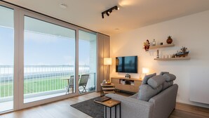 A modern living room with a large sliding glass door leading to a balcony, a grey sofa, a TV, and a small table with a lamp. - Stylish Apartment With Terrace in Bredene (Bredene)