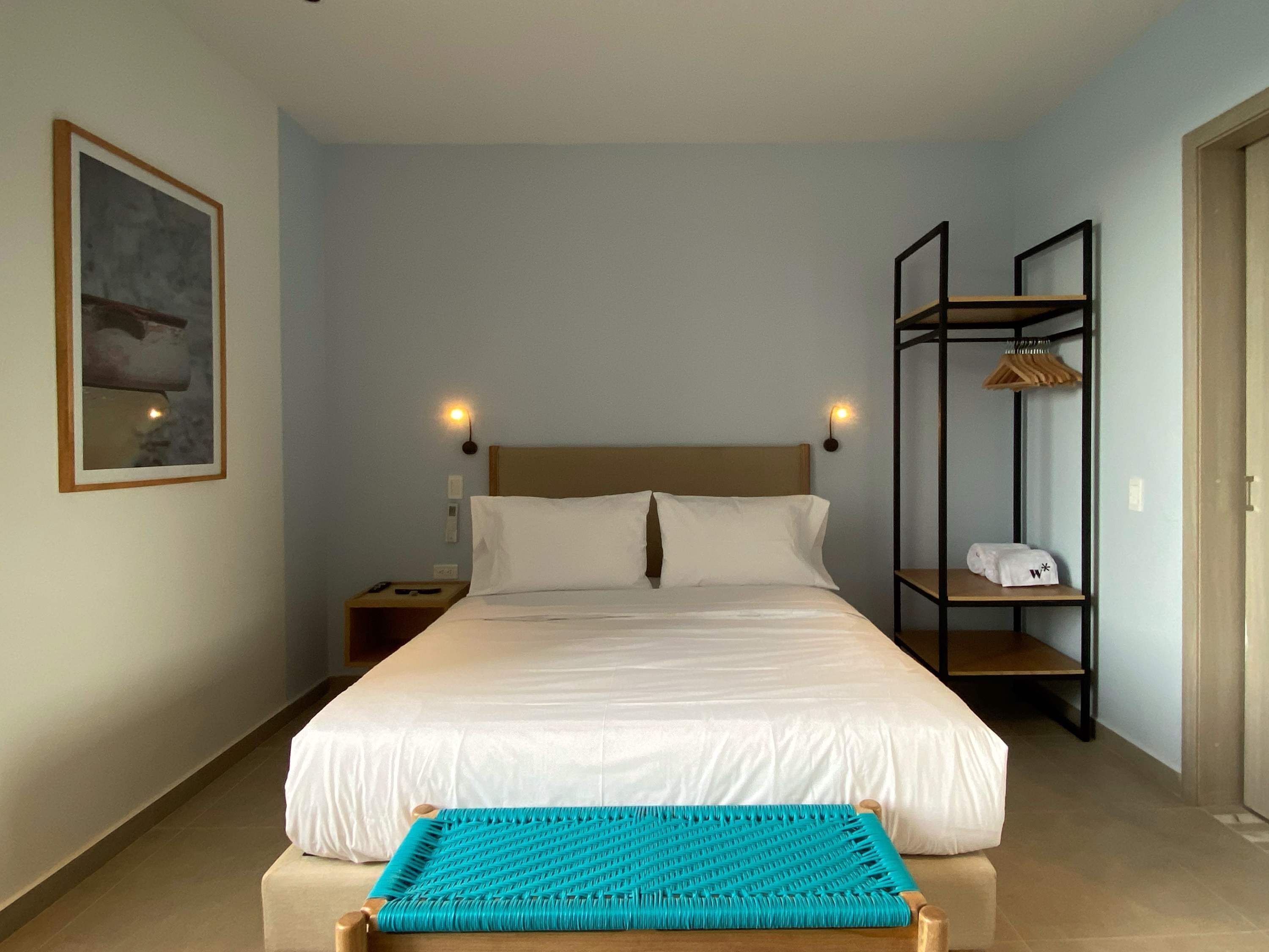 Ein ordentlich eingerichtetes Schlafzimmer mit einem Bett, einer Bank, einem Nachttisch, einem Wandregal und einem gerahmten Bild.