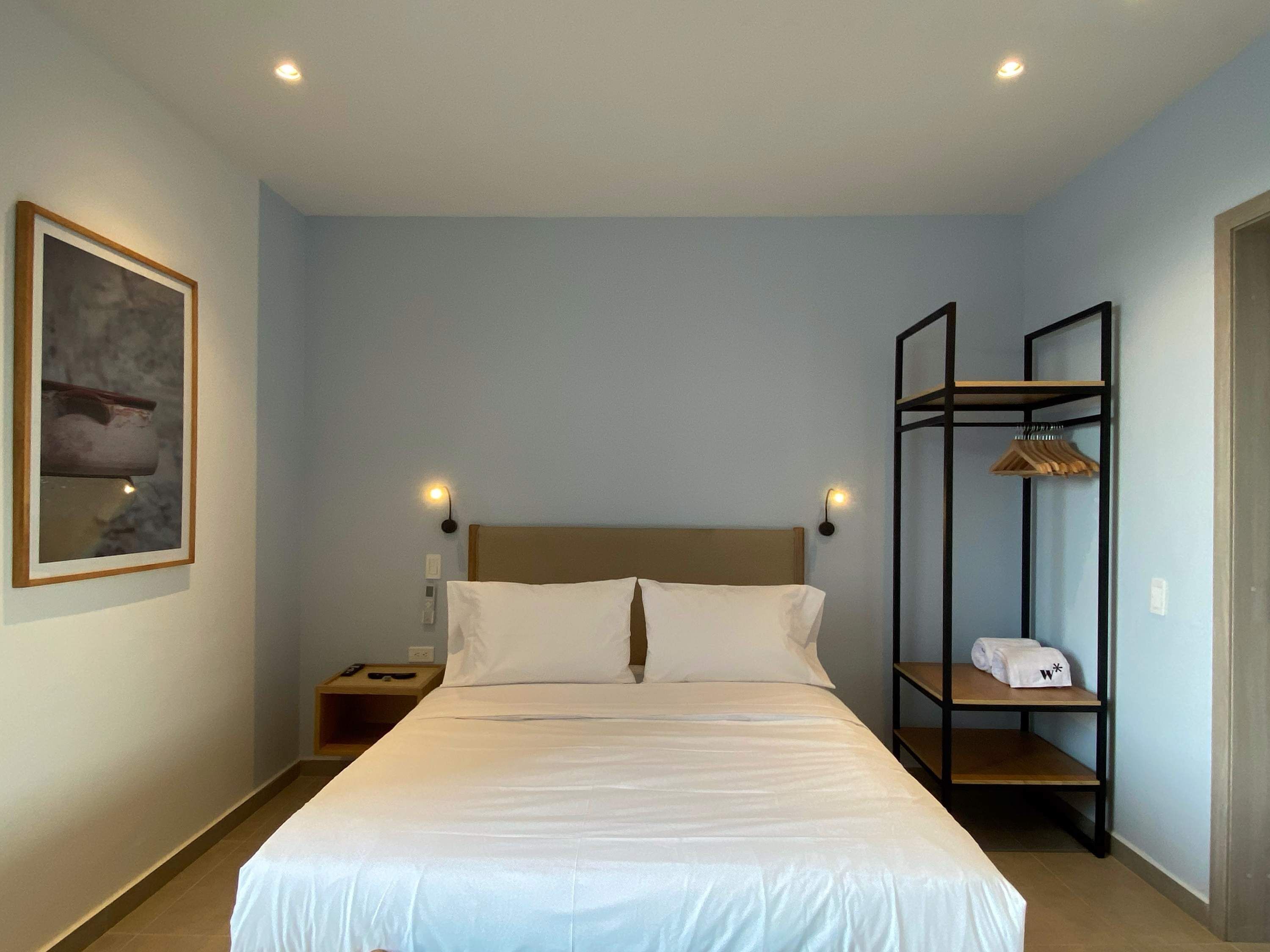 A hotel room with a large bed, two bedside tables, a wall-mounted shelf with towels, and a framed picture on the wall.