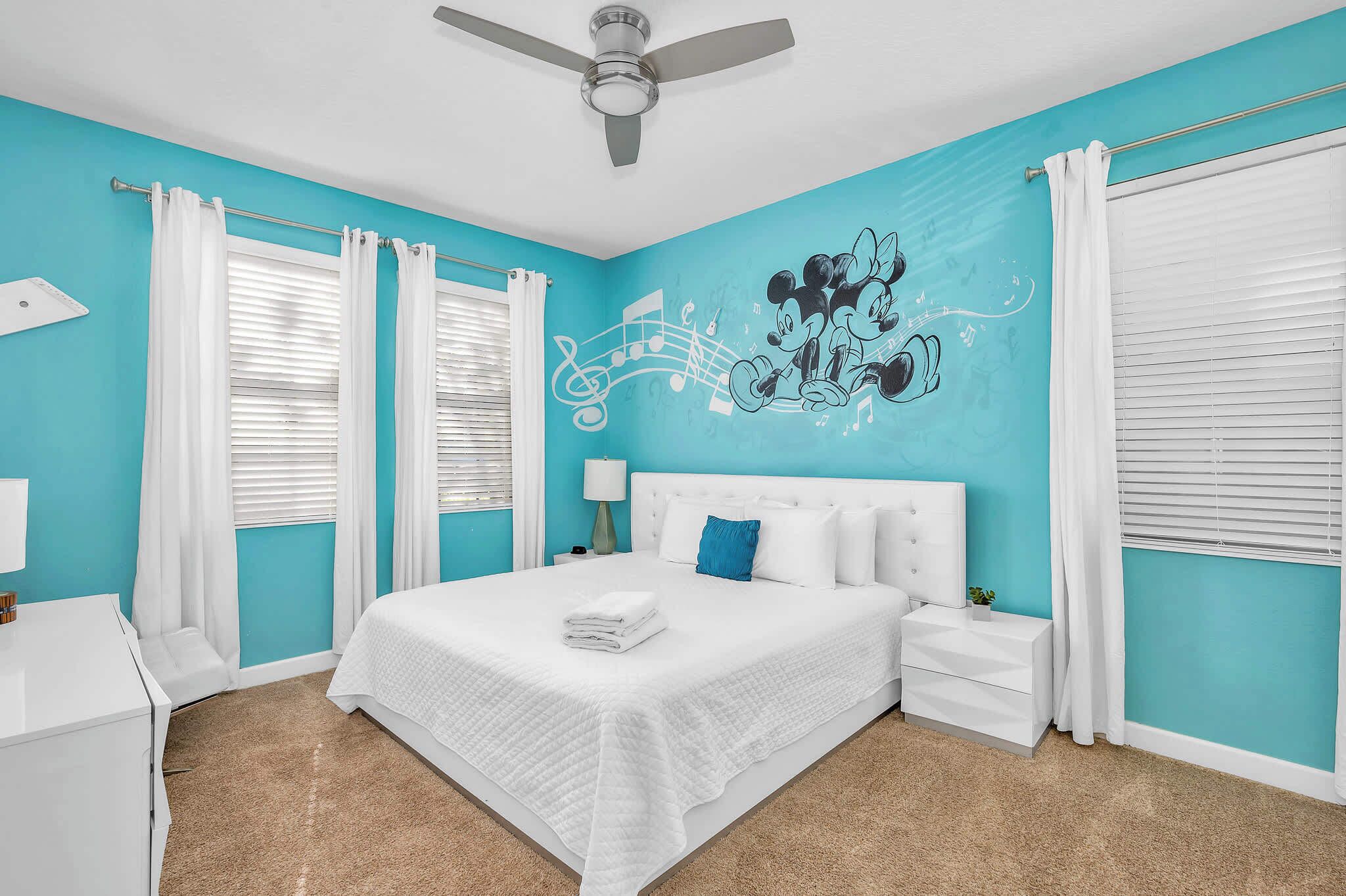 A bedroom with a bed, white curtains, a ceiling fan, and a wall mural.