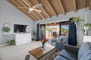 Una sala de estar con ventilador de techo, televisor, un mueble blanco, muebles con diseños y vista a un patio exterior.