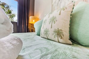 A bed with a green leaf pattern, a white pillow with palm tree designs, and a lamp in the background.